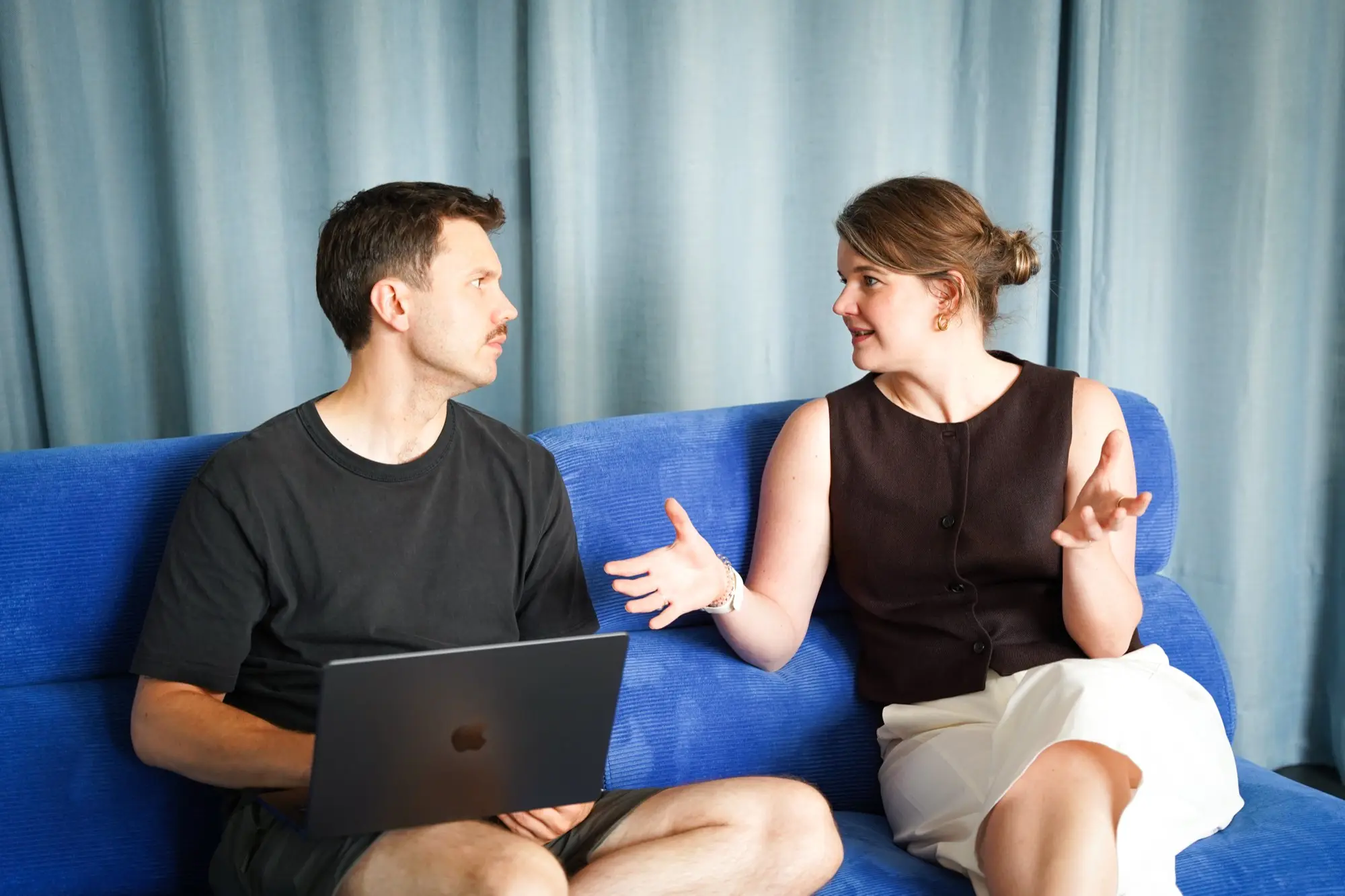 Zwei Personen im Gespräch auf einem blauen Sofa, ein Laptop aufgeklappt auf dem Schoß.